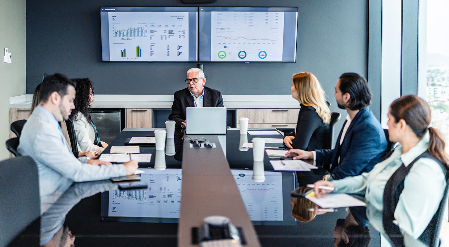 A group of individuals seated in a conference room, engaged in discussion while using a laptop for presentations.